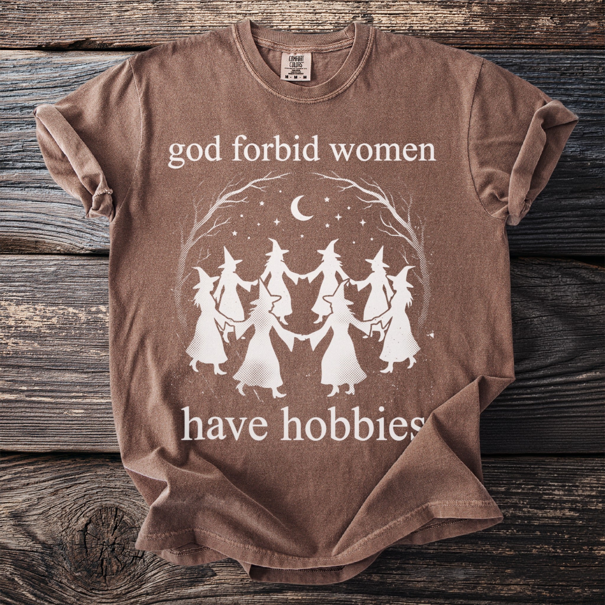 Brown t-shirt with white text that reads "god forbid women have hobbies" and a graphic of witches dancing.