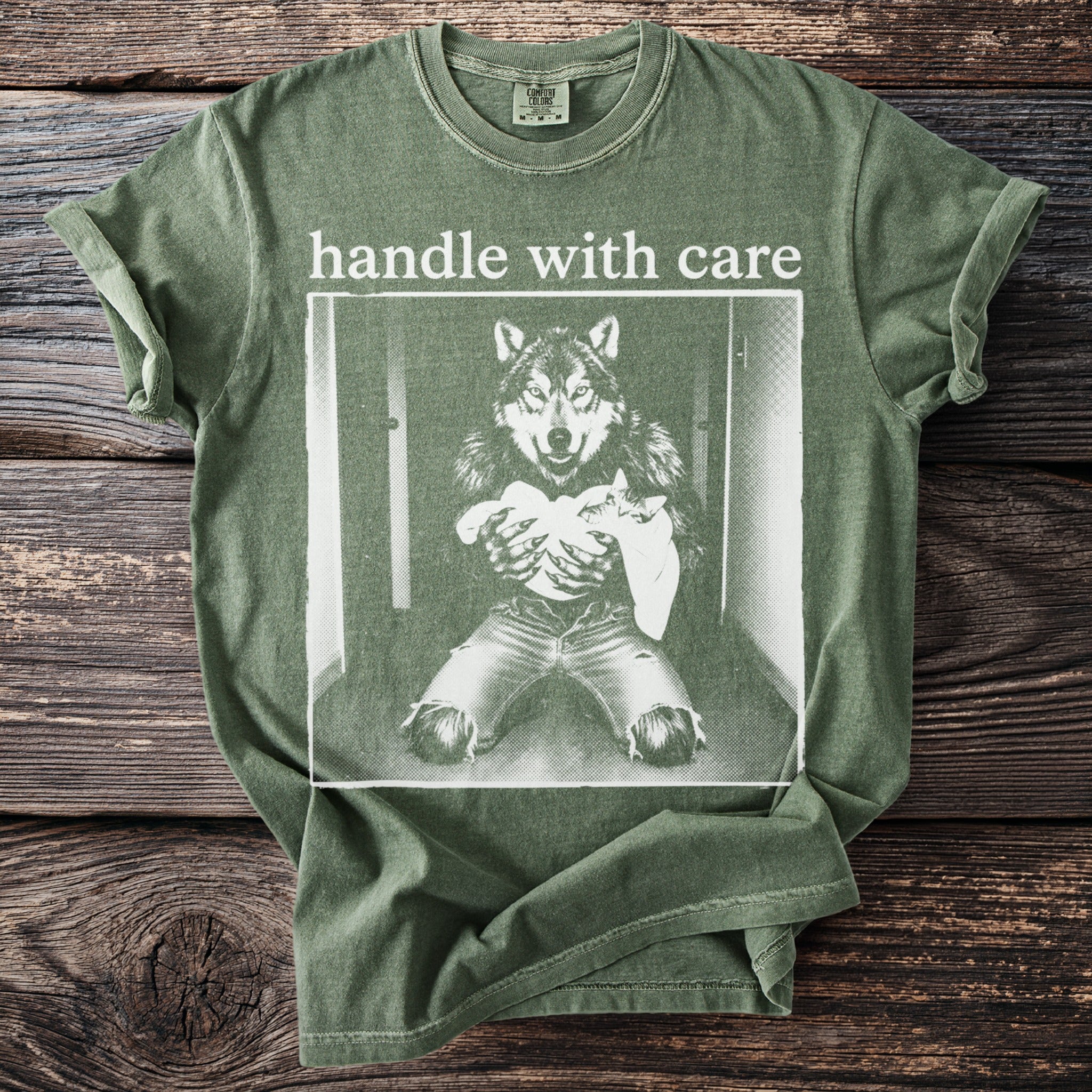 Green t-shirt with graphic of a wolf holding a baby, text reads "handle with care".