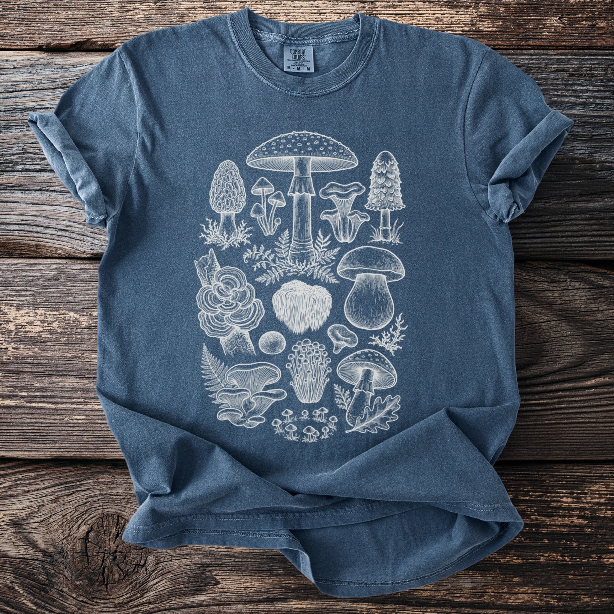 Blue t-shirt with white line art of various mushrooms and ferns on a wooden background.