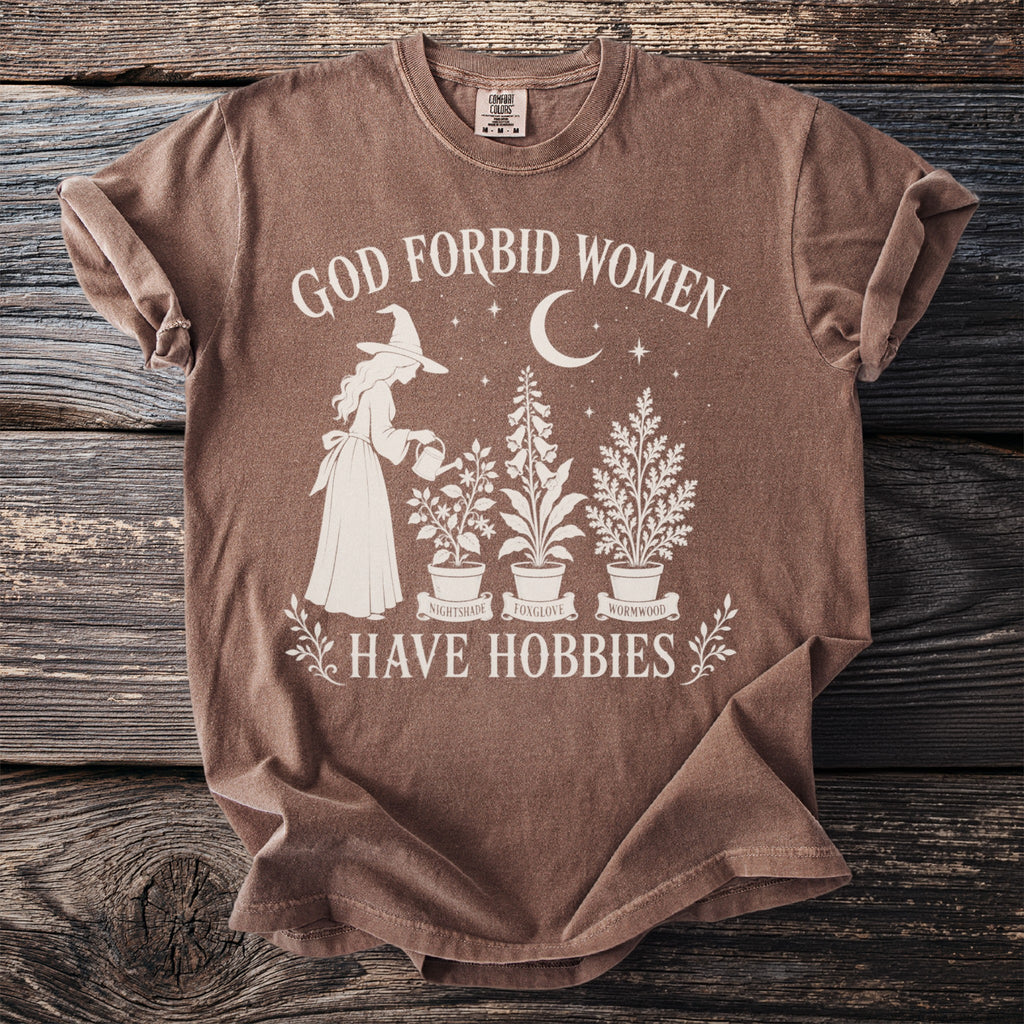 Brown t-shirt with graphic reading "God Forbid Women Have Hobbies" and witch watering plants.