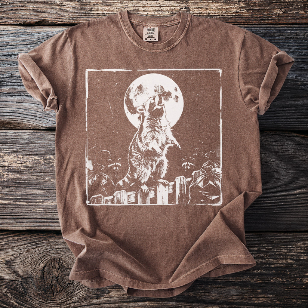 Brown t-shirt with a white graphic of a raccoon howling at the moon over trash bags on a fence.
