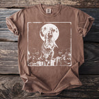 Brown t-shirt with a white graphic of a raccoon howling at the moon over trash bags on a fence.