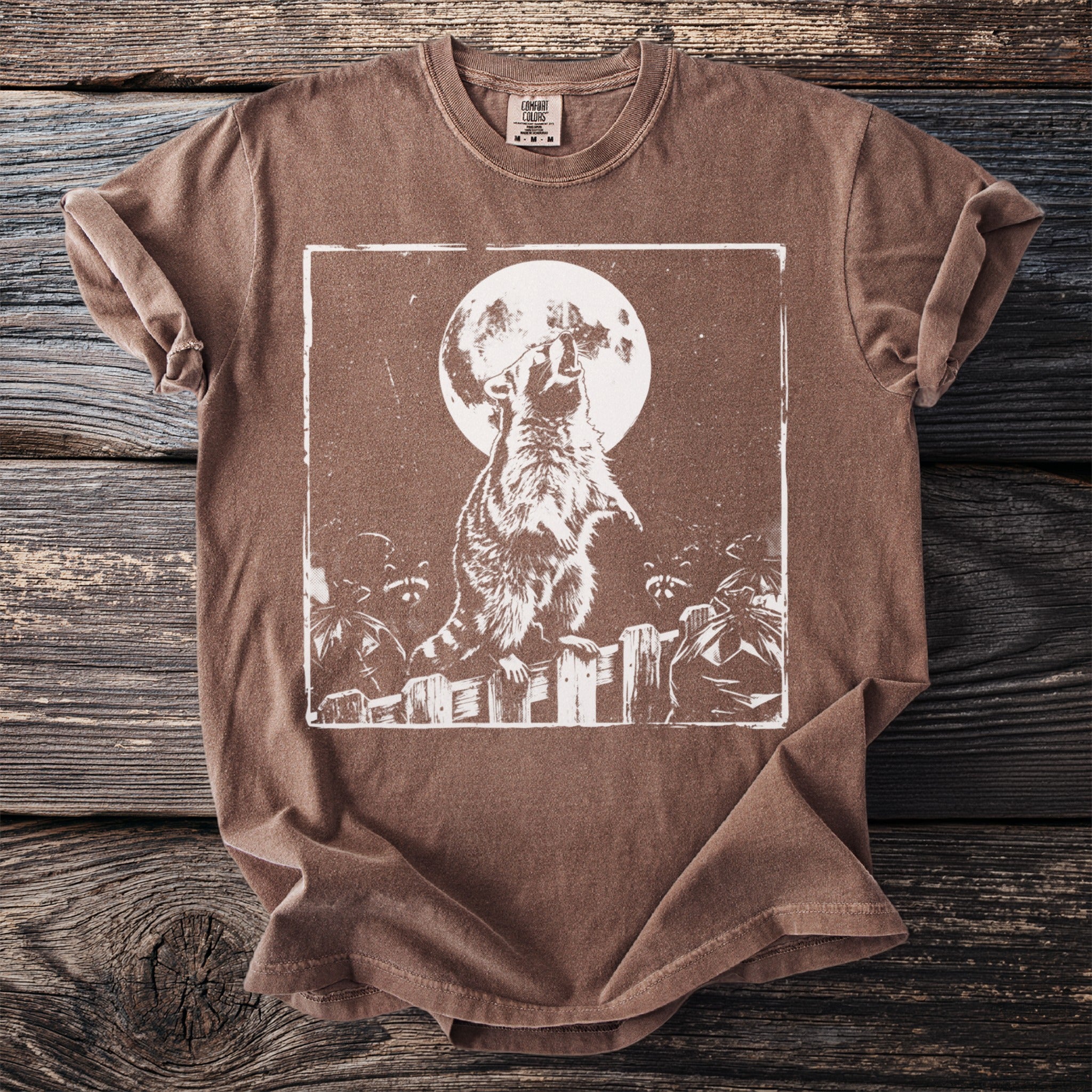 Brown t-shirt with a white graphic of a raccoon howling at the moon over trash bags on a fence.