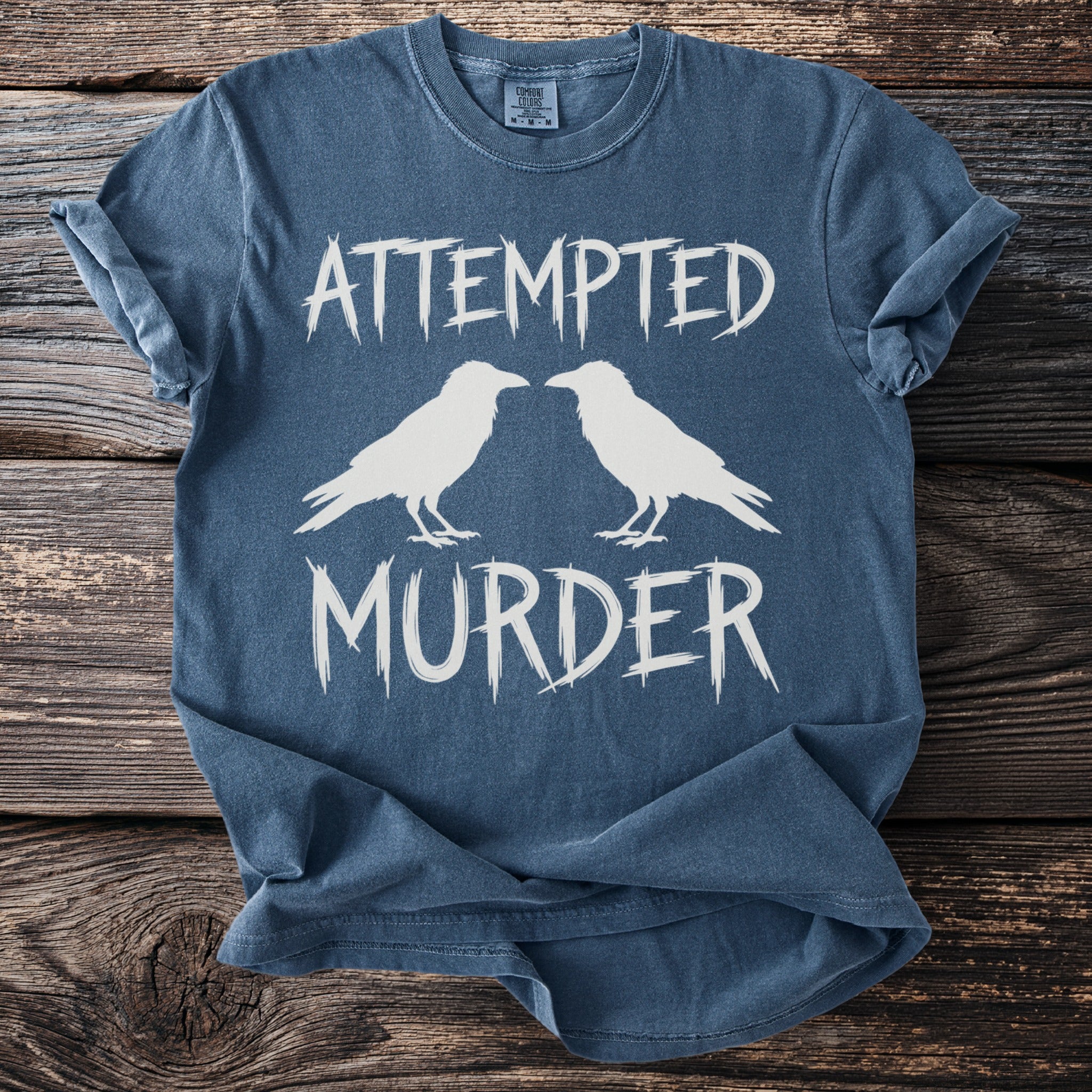 Blue t-shirt with white text "ATTEMPTED MURDER" and two white raven silhouettes.