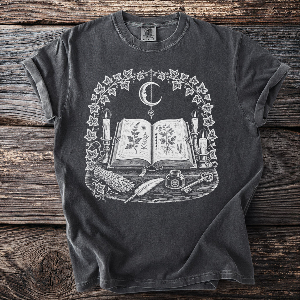 Dark gray t-shirt with a white occult design featuring an open book, moon, candles, and herbs.