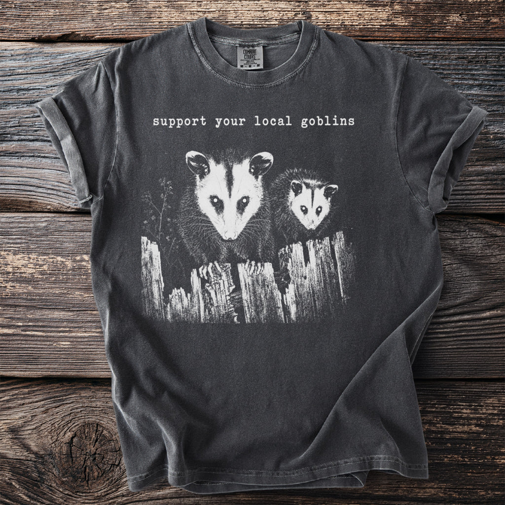 Dark t-shirt graphic with two opossums on a fence, text reads "support your local goblins."