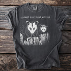 Dark t-shirt graphic with two opossums on a fence, text reads "support your local goblins."