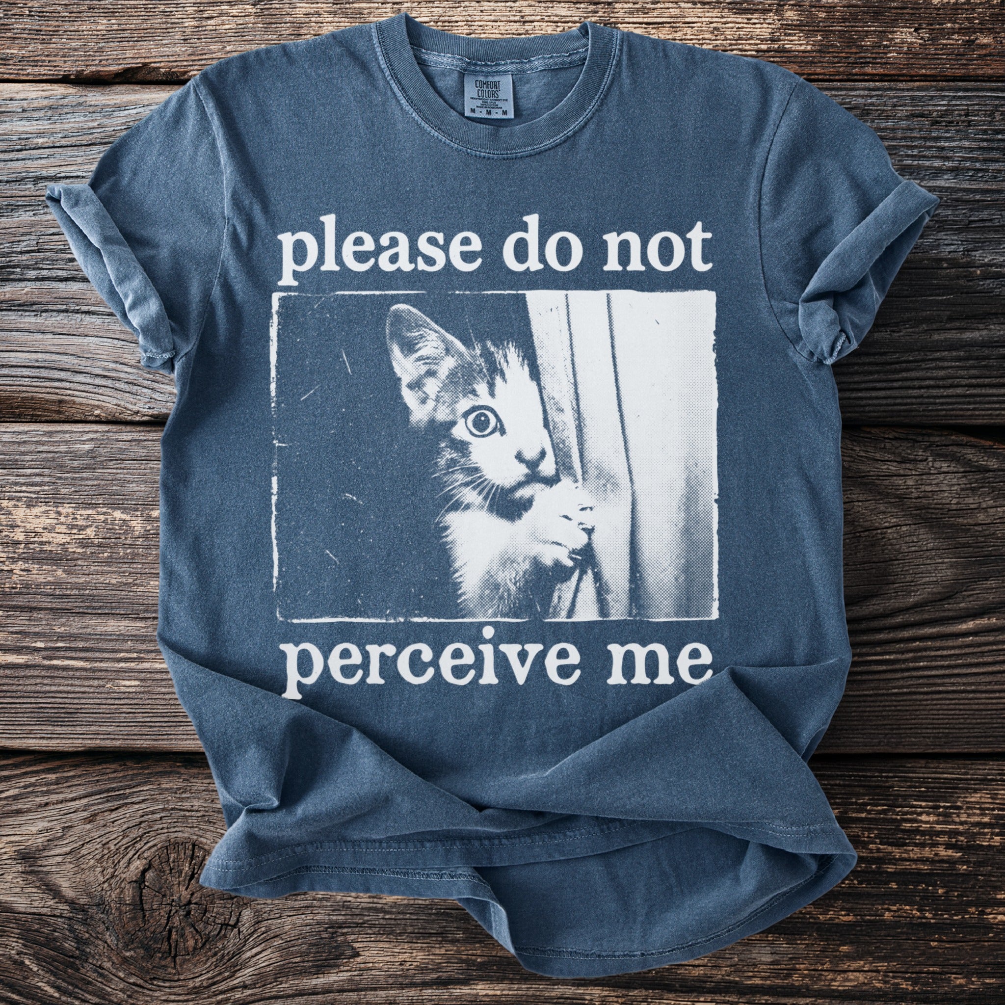 Blue t-shirt with a kitten peeking from behind a curtain, text says "please do not perceive me."