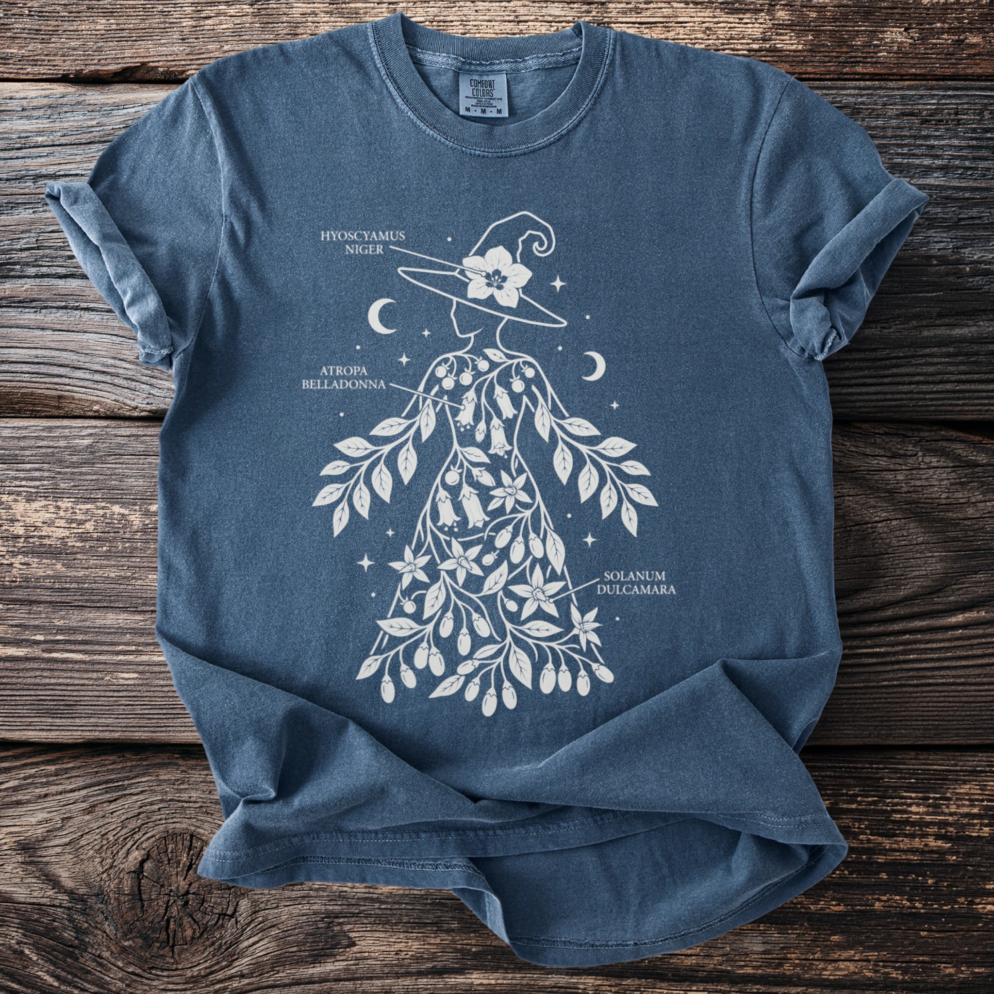 Blue t-shirt with witch design featuring botanical illustrations and text labels.