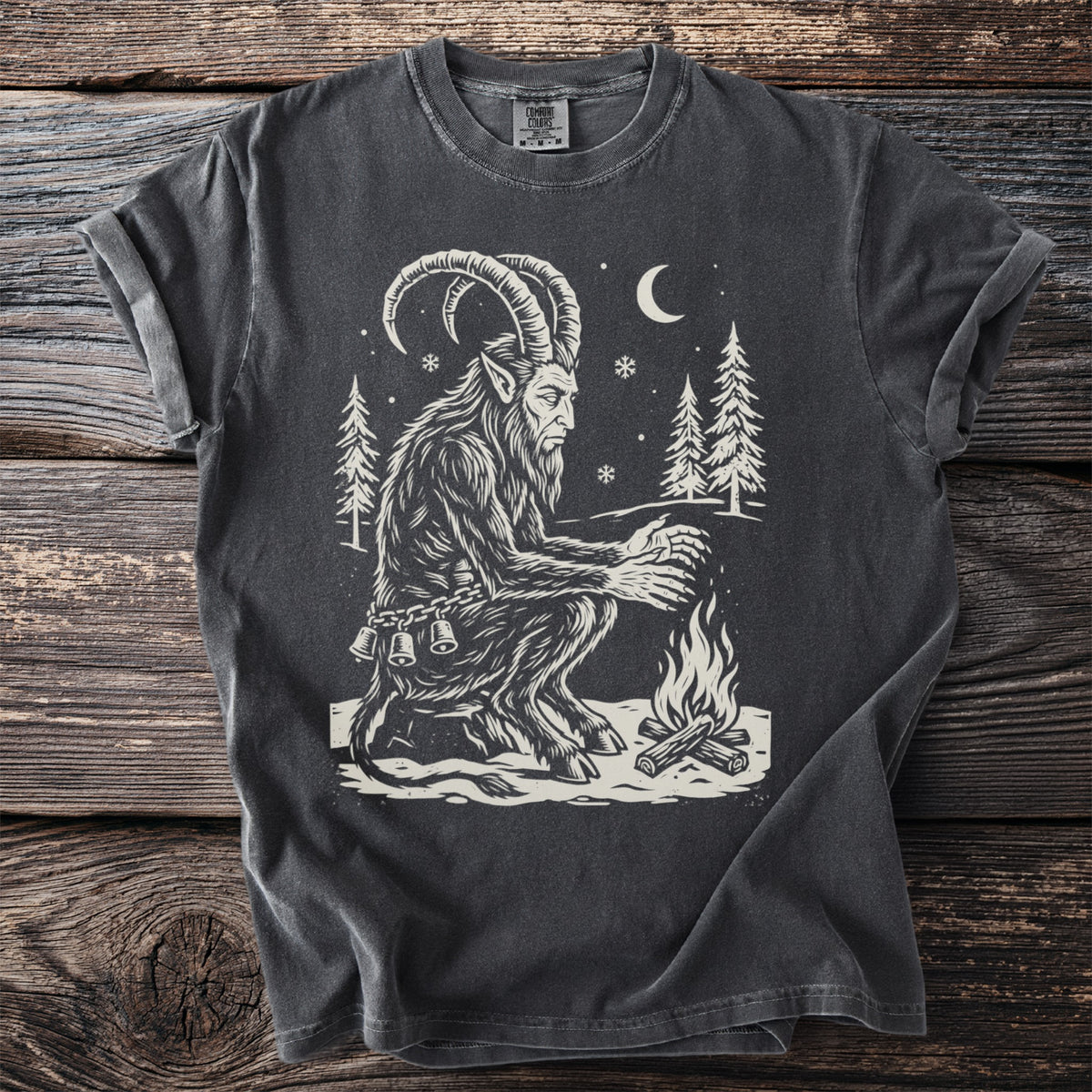 Pepper gray t-shirt with a cream folk-cut print: Krampus sits in the snow among tall pines, warming his clawed hands over a small fire.