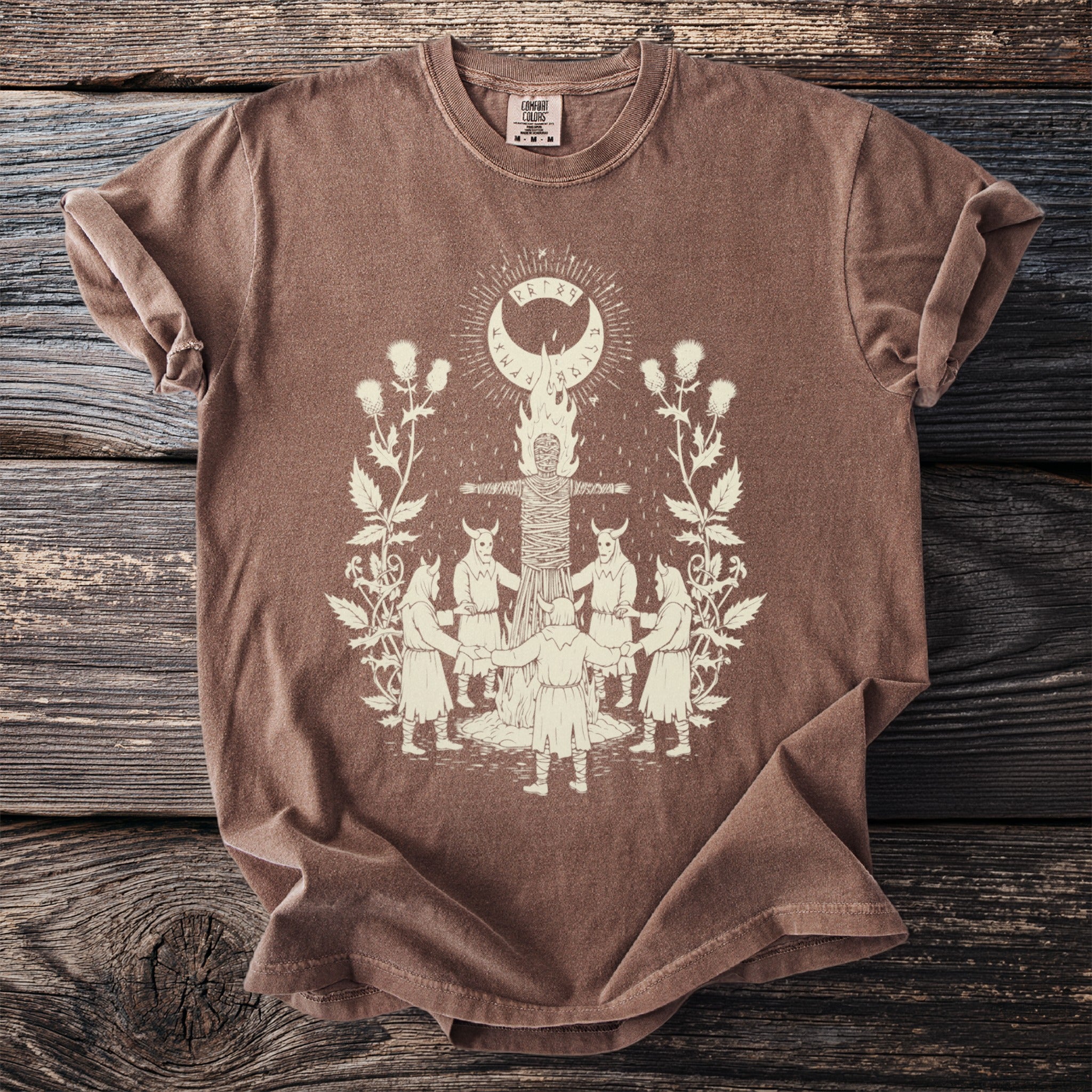 Brown t-shirt with a cream graphic of a ritualistic scene with people and a figure on a pyre.