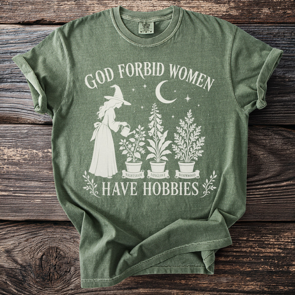 Green t-shirt with white graphic of a witch watering plants, text "GOD FORBID WOMEN HAVE HOBBIES".