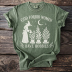 Green t-shirt with white graphic of a witch watering plants, text "GOD FORBID WOMEN HAVE HOBBIES".
