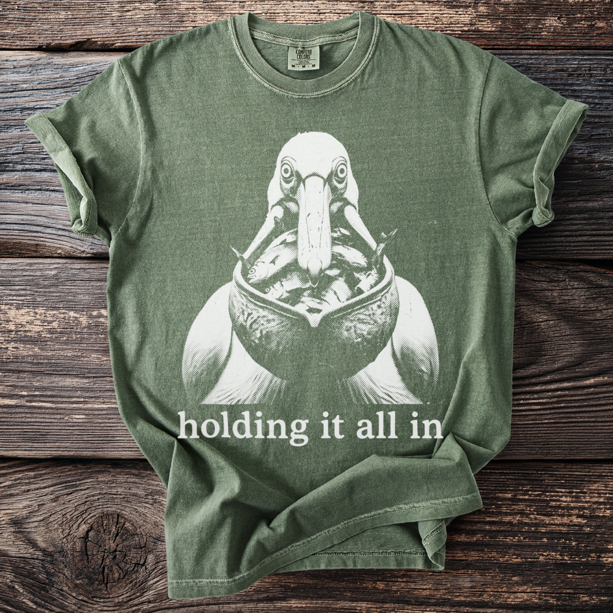 Green t-shirt with a pelican graphic holding many fish and text "holding it all in."