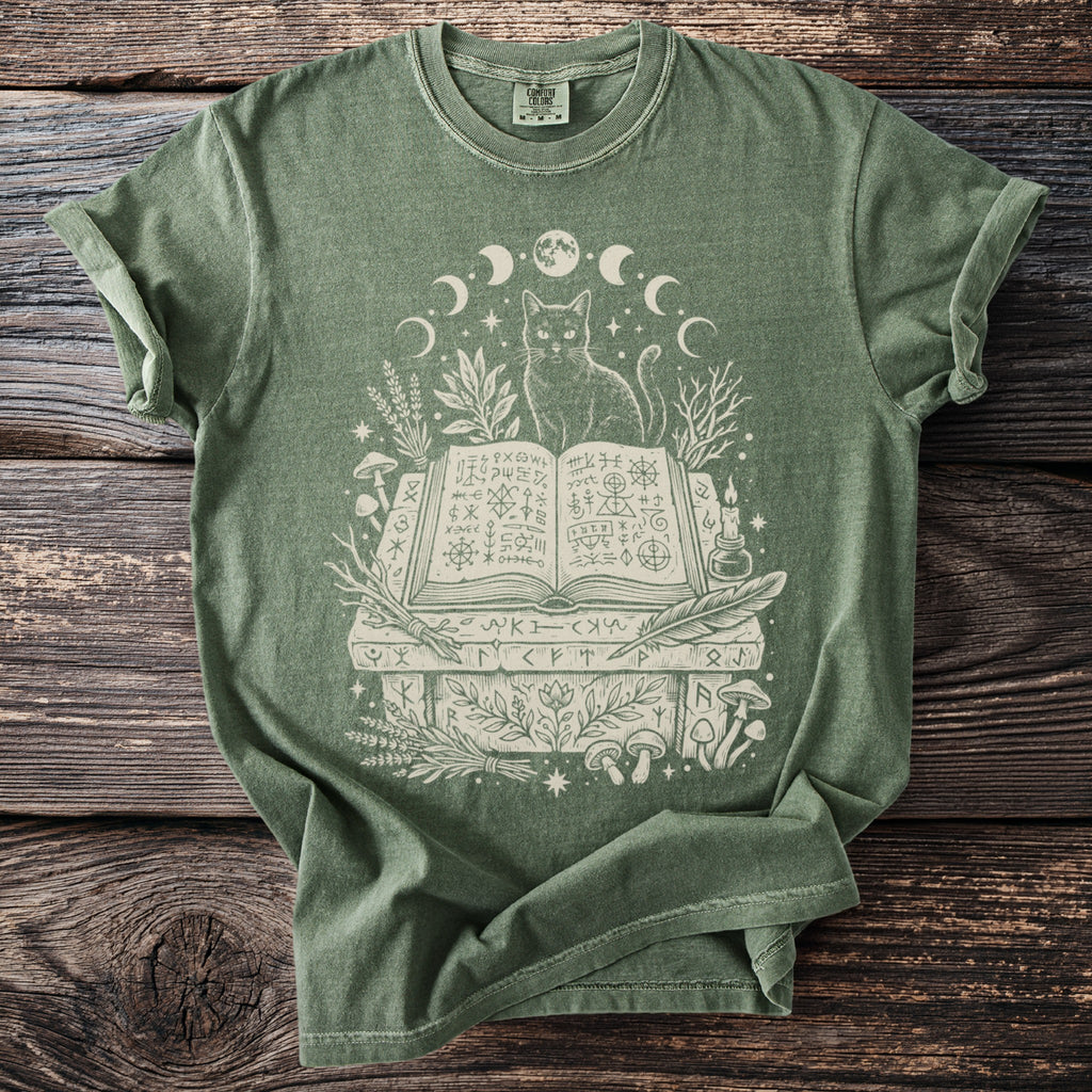 Green t-shirt with white graphic of a cat, open book, moons, plants, and mushrooms.