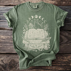 Green t-shirt with white graphic of a cat, open book, moons, plants, and mushrooms.