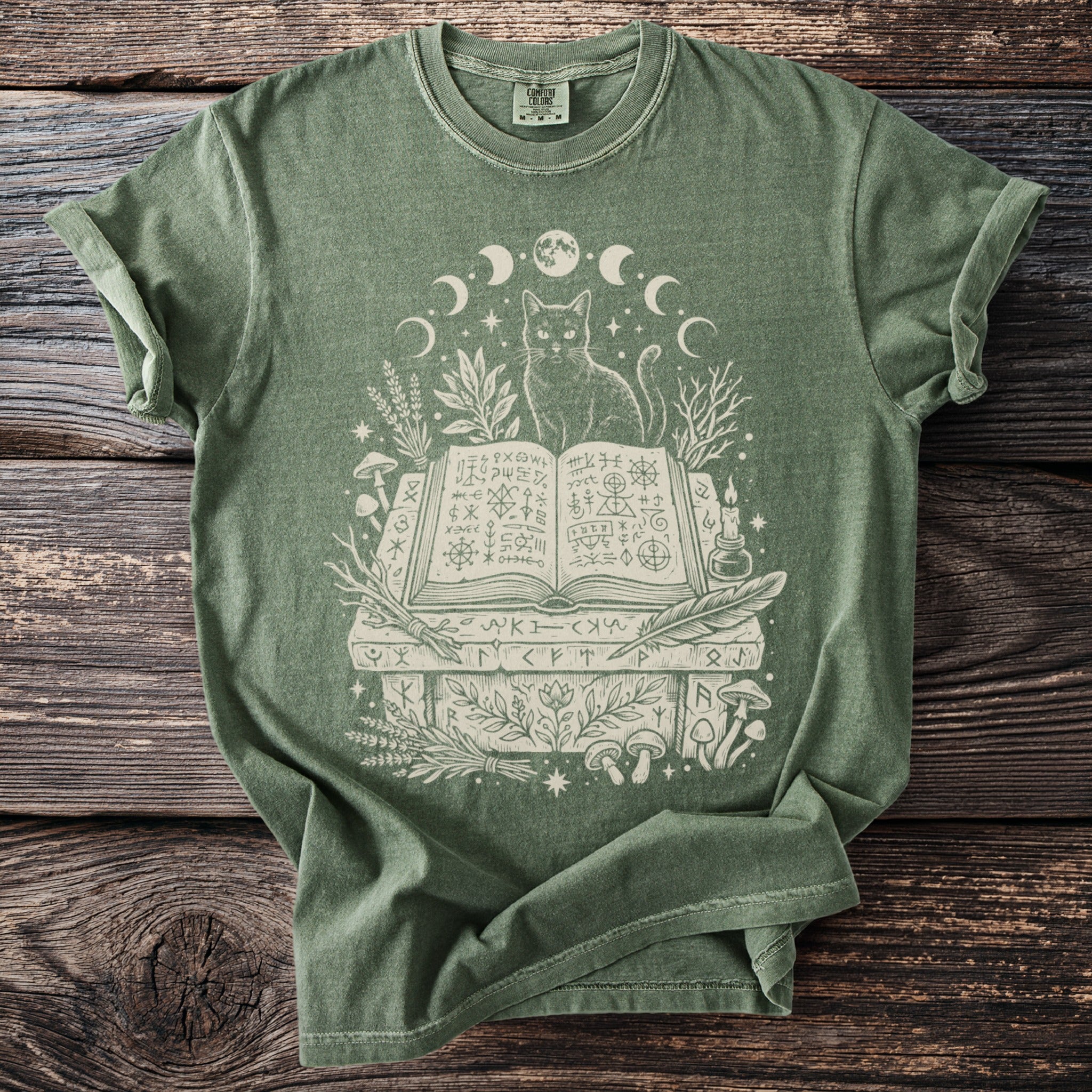 Green t-shirt with white graphic of a cat, open book, moons, plants, and mushrooms.