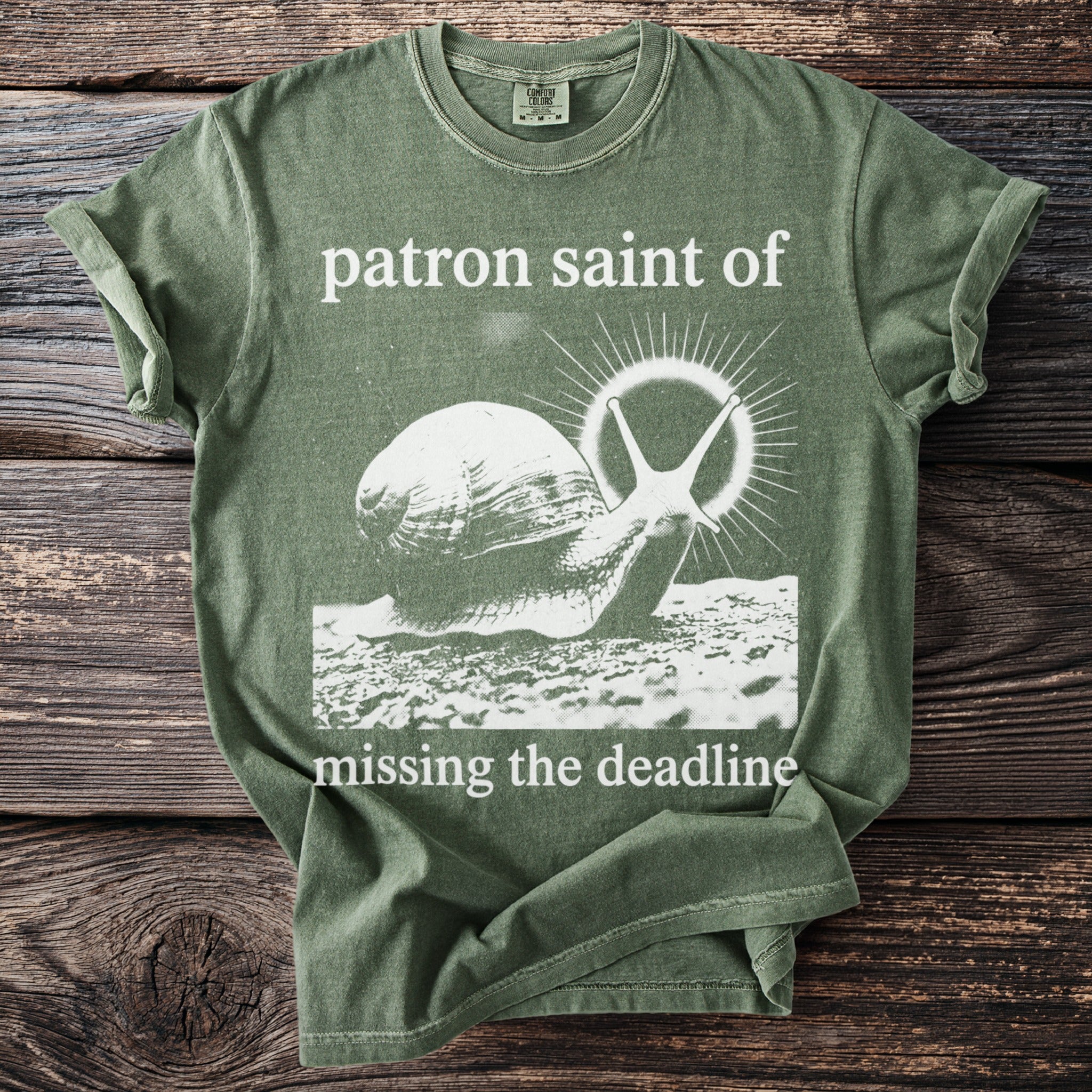Olive green t-shirt with a snail graphic and text "patron saint of missing the deadline".