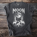 Dark grey t-shirt graphic with white text "MOON", a full moon, two towers, a dog, a wolf, a river, and a lobster.