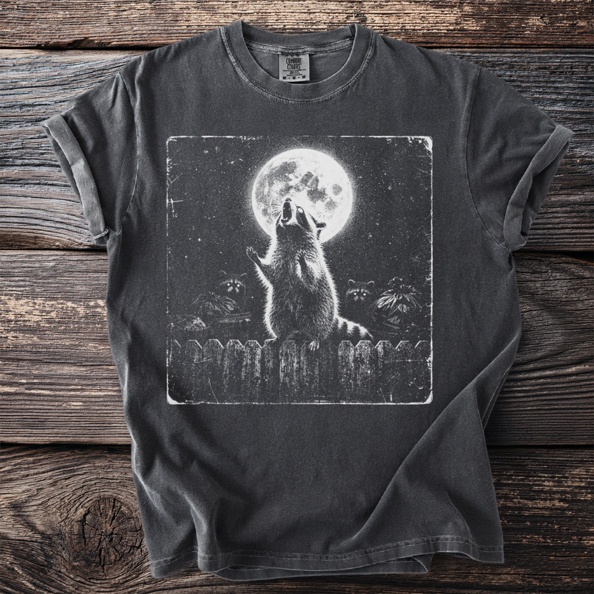 Graphic t-shirt with raccoon howling at full moon over trash bins and fence.