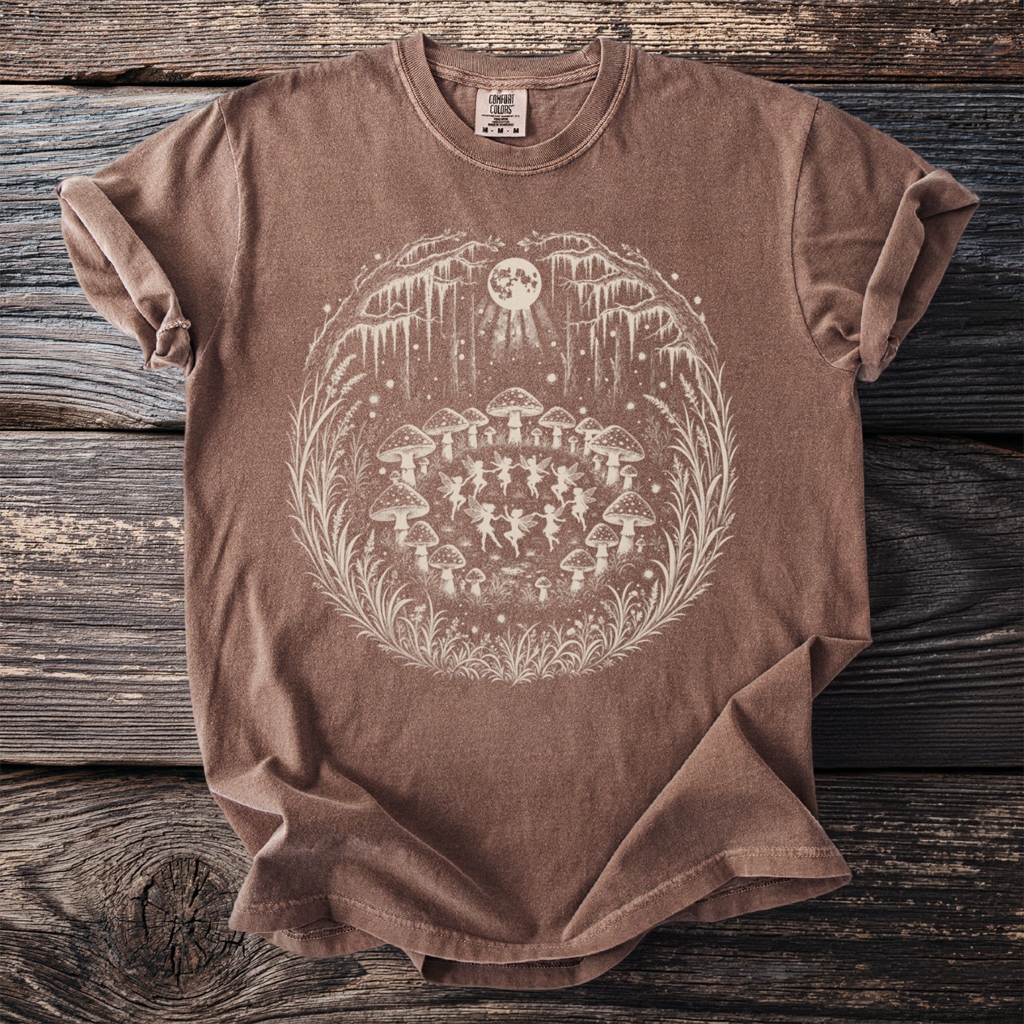 Brown t-shirt with a circular graphic of fairies dancing among mushrooms under a full moon.
