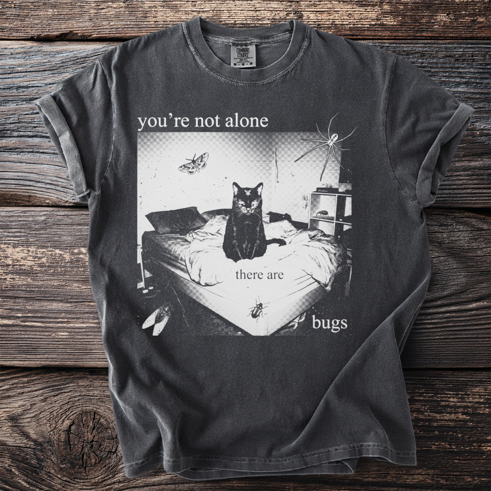 Black t-shirt with graphic of cat on a bed with butterflies, spiders, and bugs, "you're not alone there are bugs."