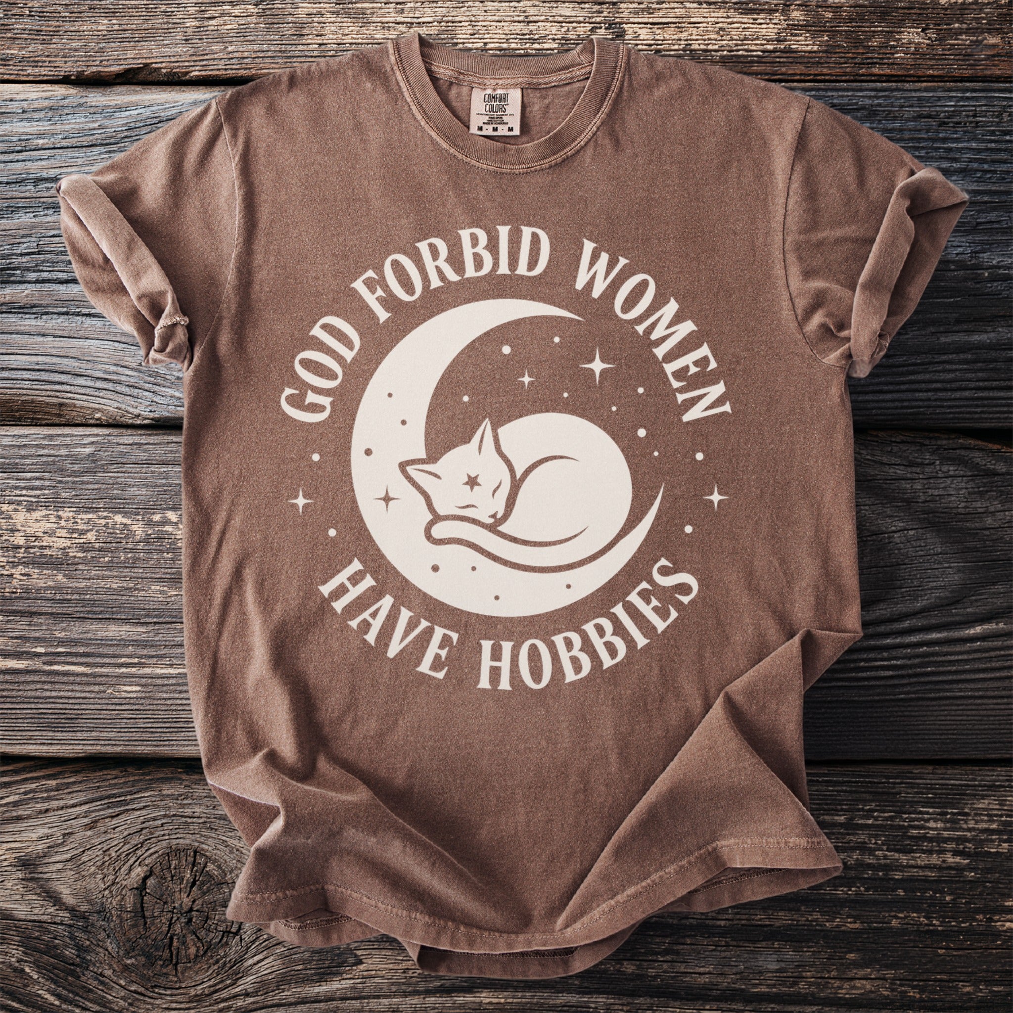 Brown t-shirt with "God Forbid Women Have Hobbies" text and a sleeping cat on a moon.