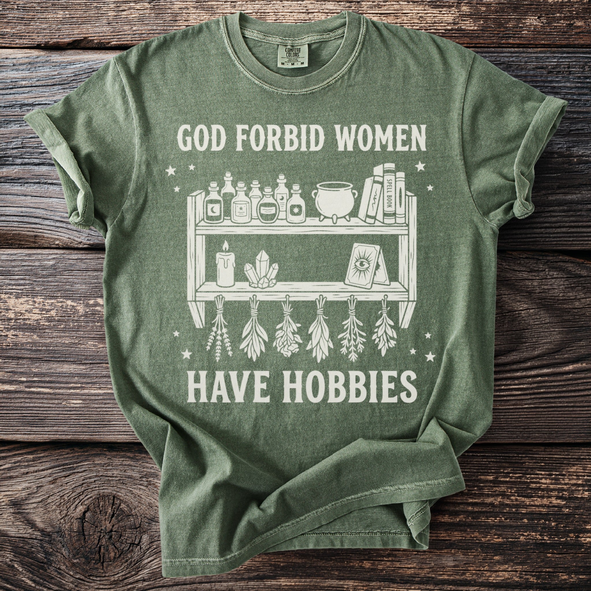 Green t-shirt with white graphic text "GOD FORBID WOMEN HAVE HOBBIES" and witchy imagery.