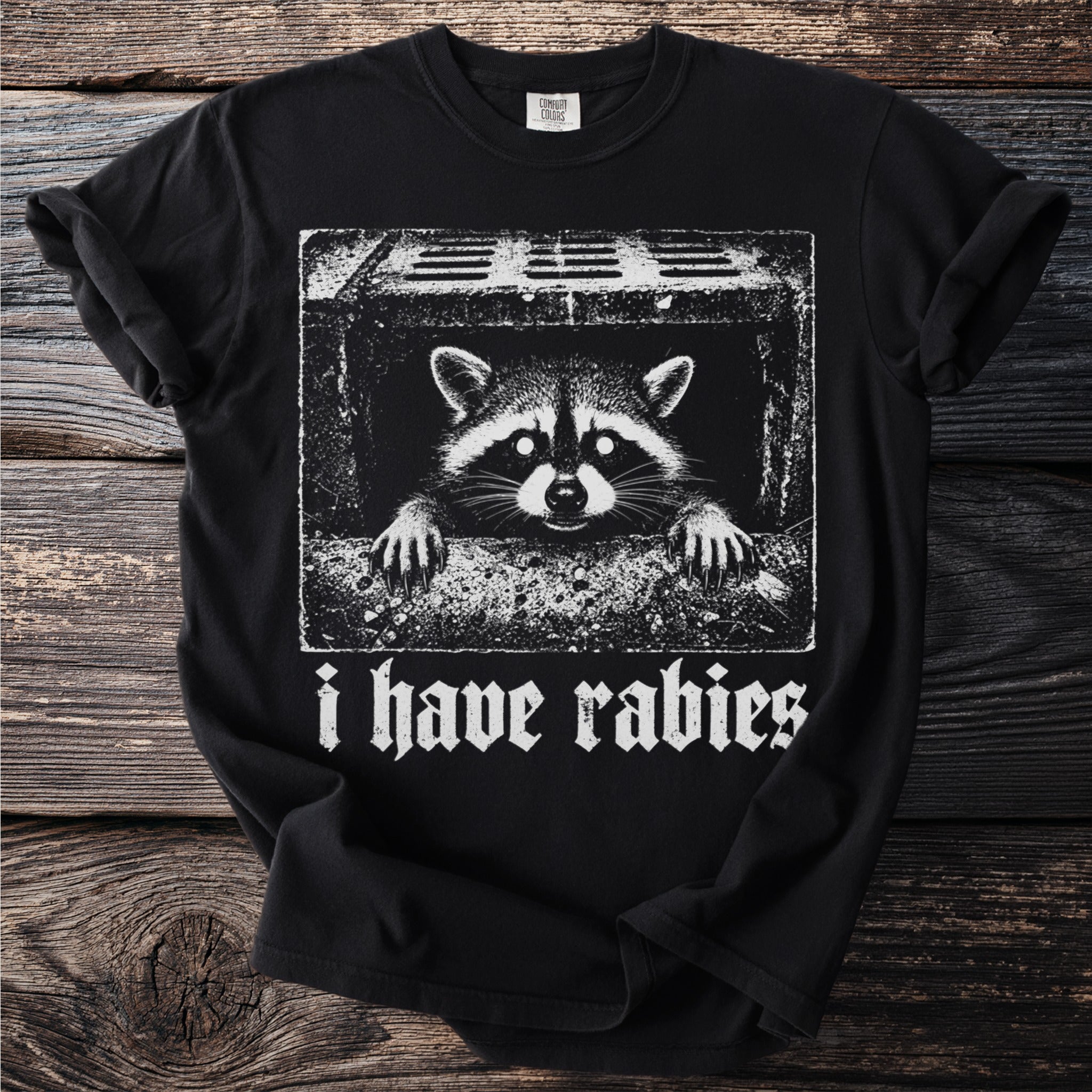 Black t-shirt with a stark white graphic of a raccoon peeking from a drainpipe, text reads "i have rabies".