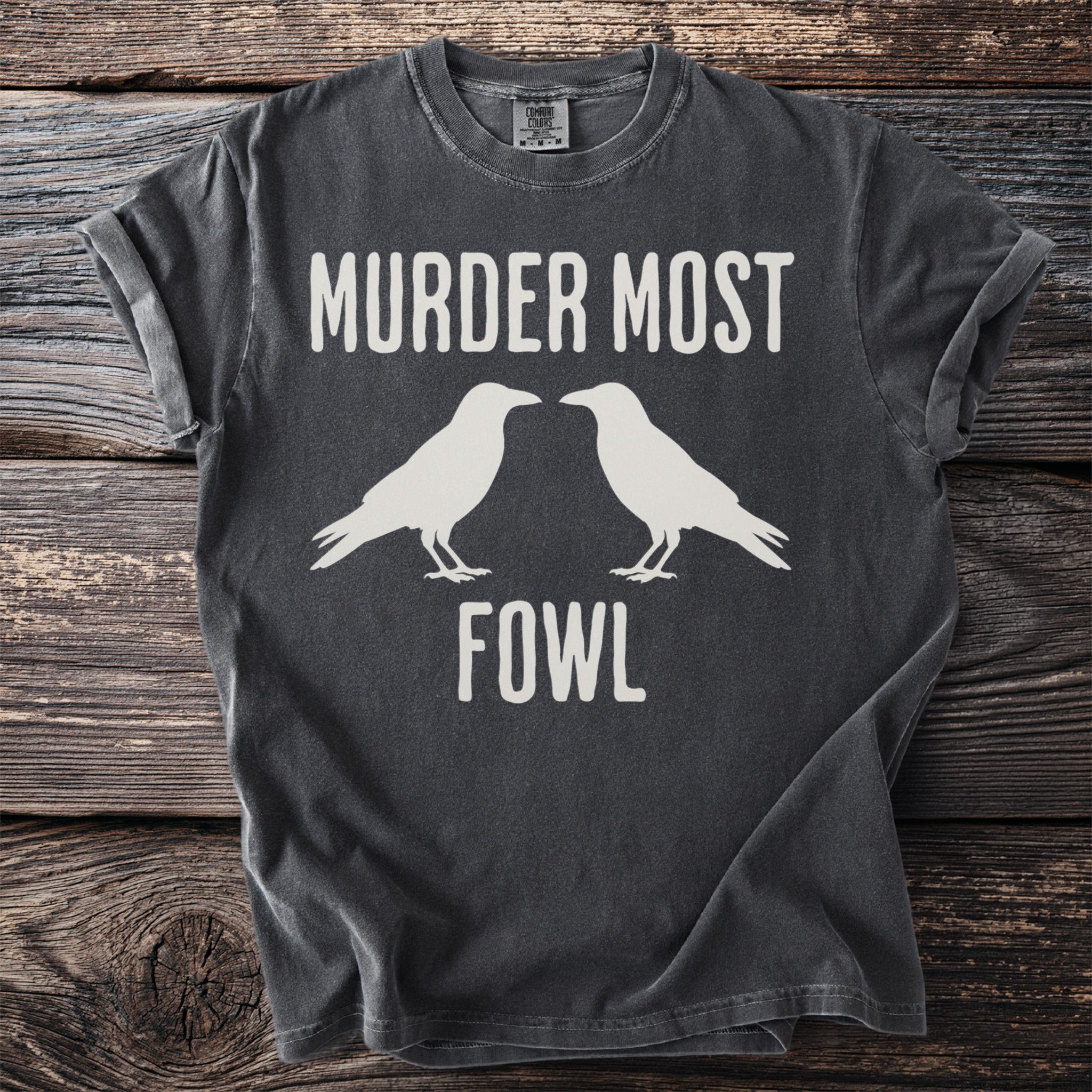 Dark gray t-shirt with white text "MURDER MOST FOWL" and two crow silhouettes.