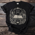 Black t-shirt graphic with black cat, open book, candles, mushrooms, and moons.