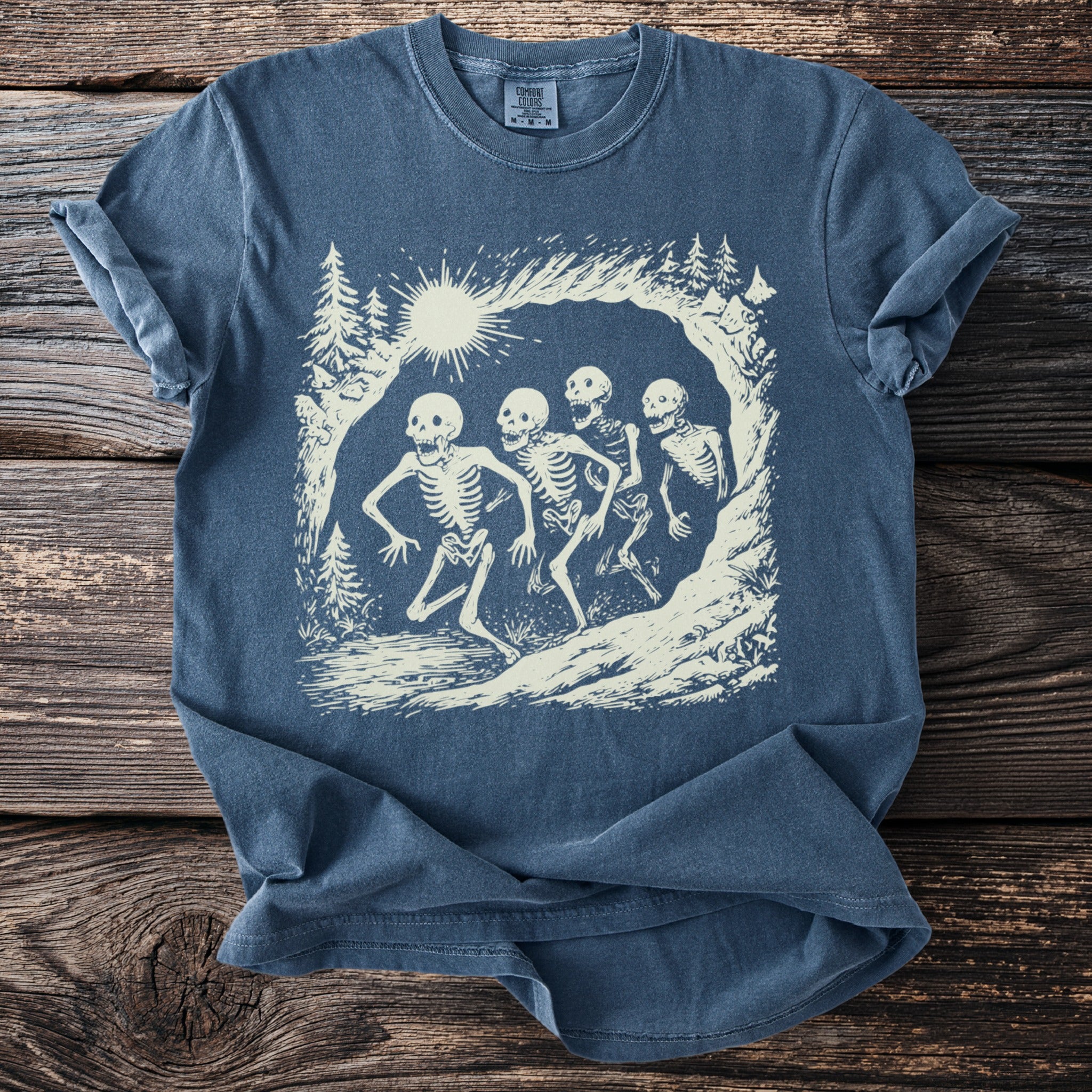 Blue t-shirt with a graphic of four skeletons running in a forest landscape.