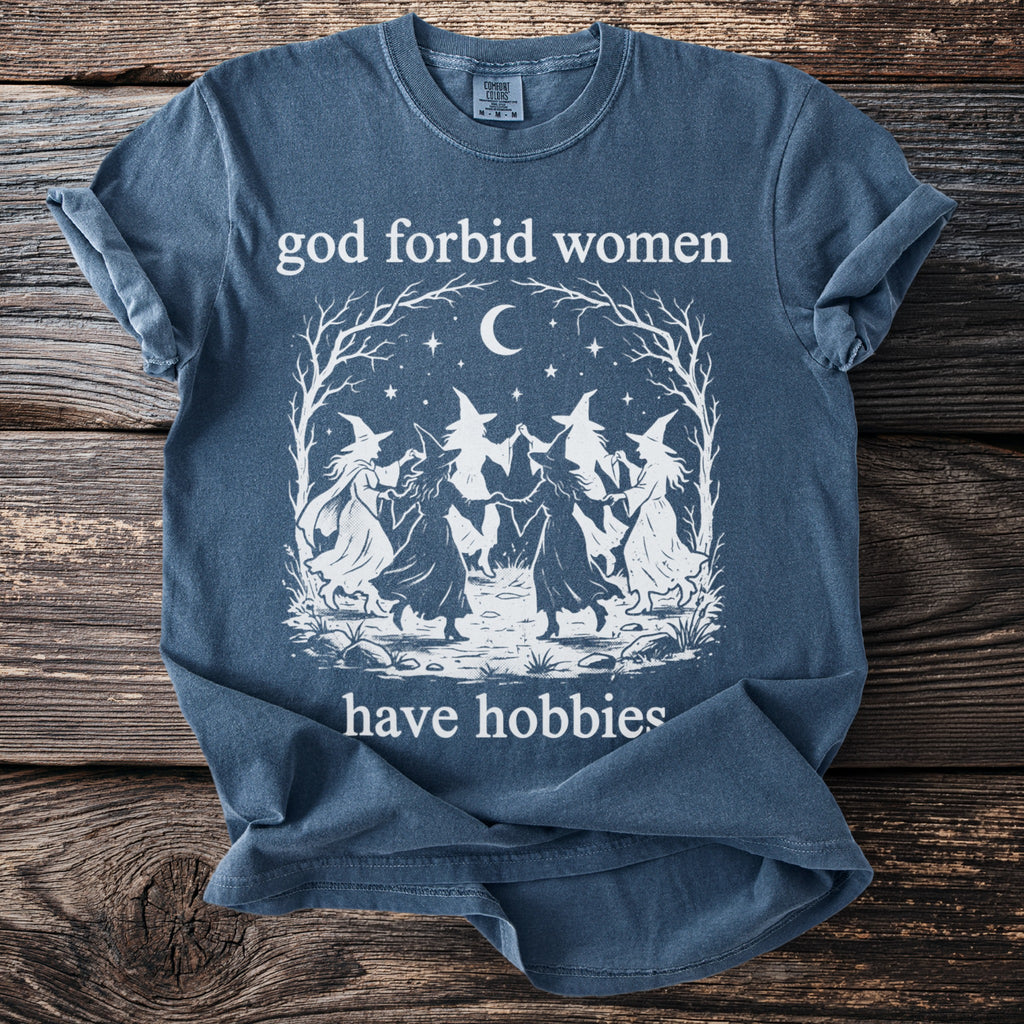 Blue t-shirt with white graphic of witches dancing under a moon and text "god forbid women have hobbies".