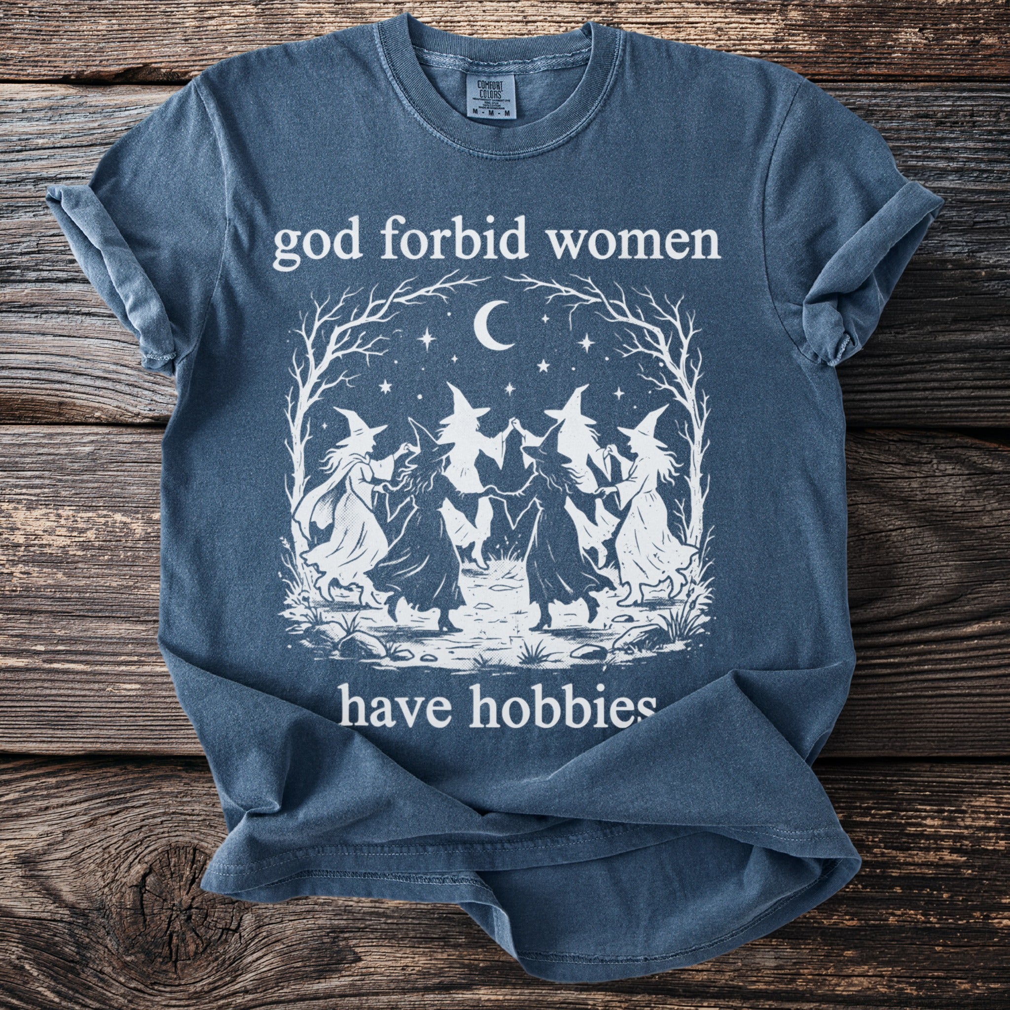 Blue t-shirt with white graphic of witches dancing under a moon and text "god forbid women have hobbies".