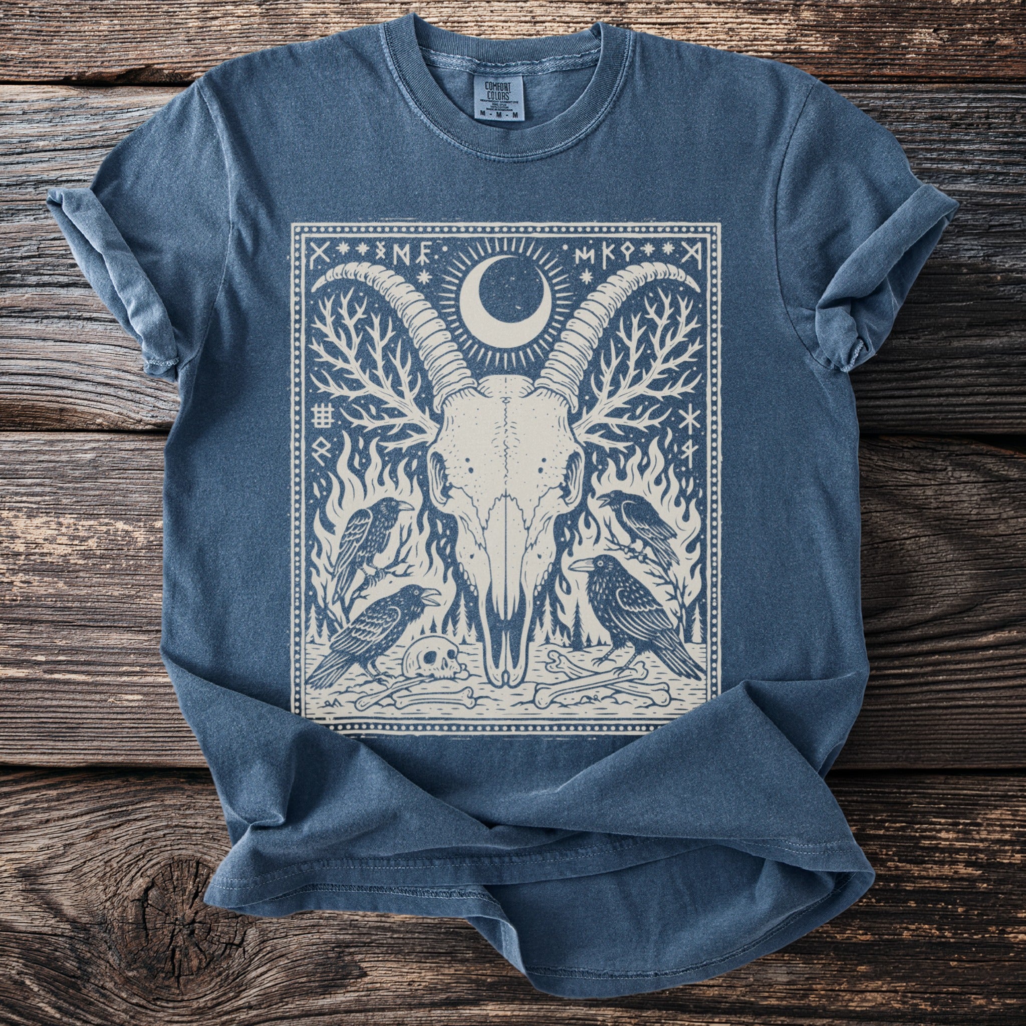 Blue t-shirt with a cream graphic of a goat skull with moon, ravens, bones, and flames.