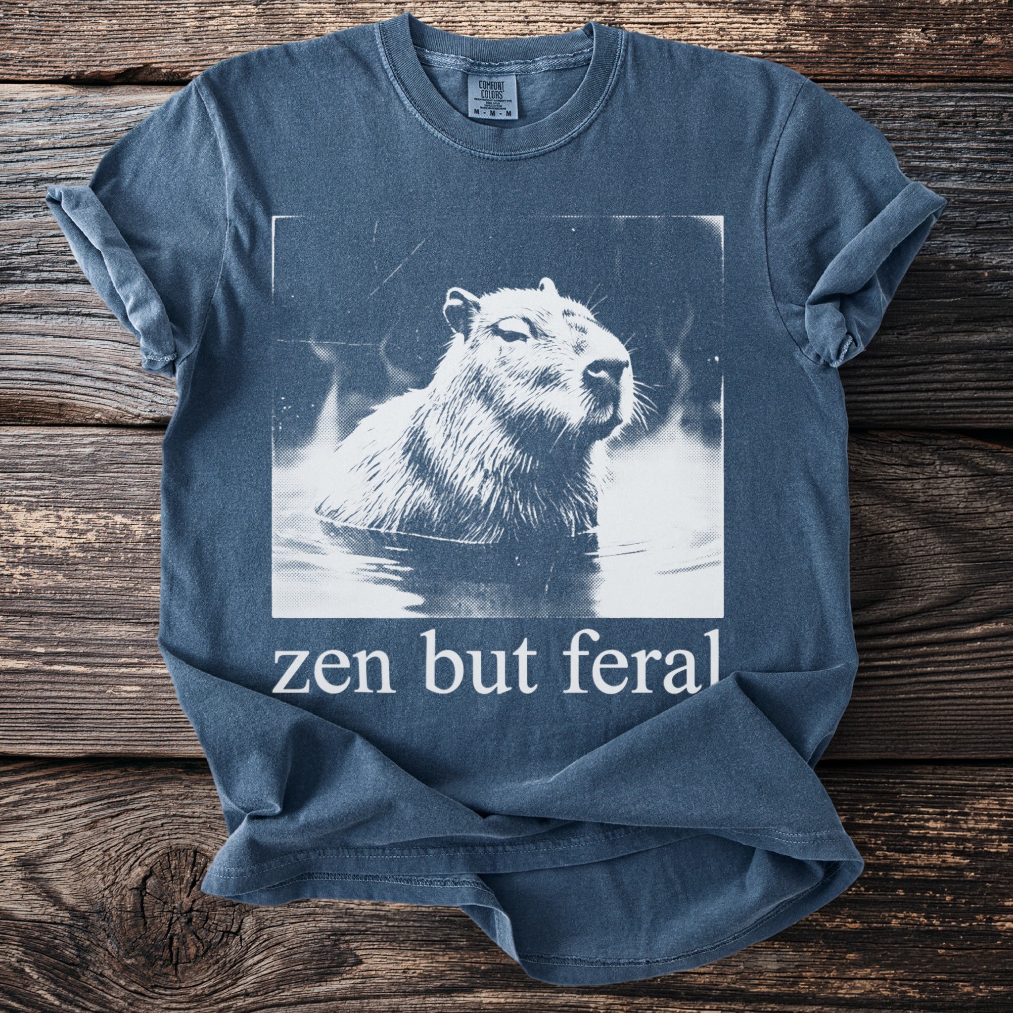 Capybara in hot spring graphic with "zen but feral" text on a blue t-shirt.