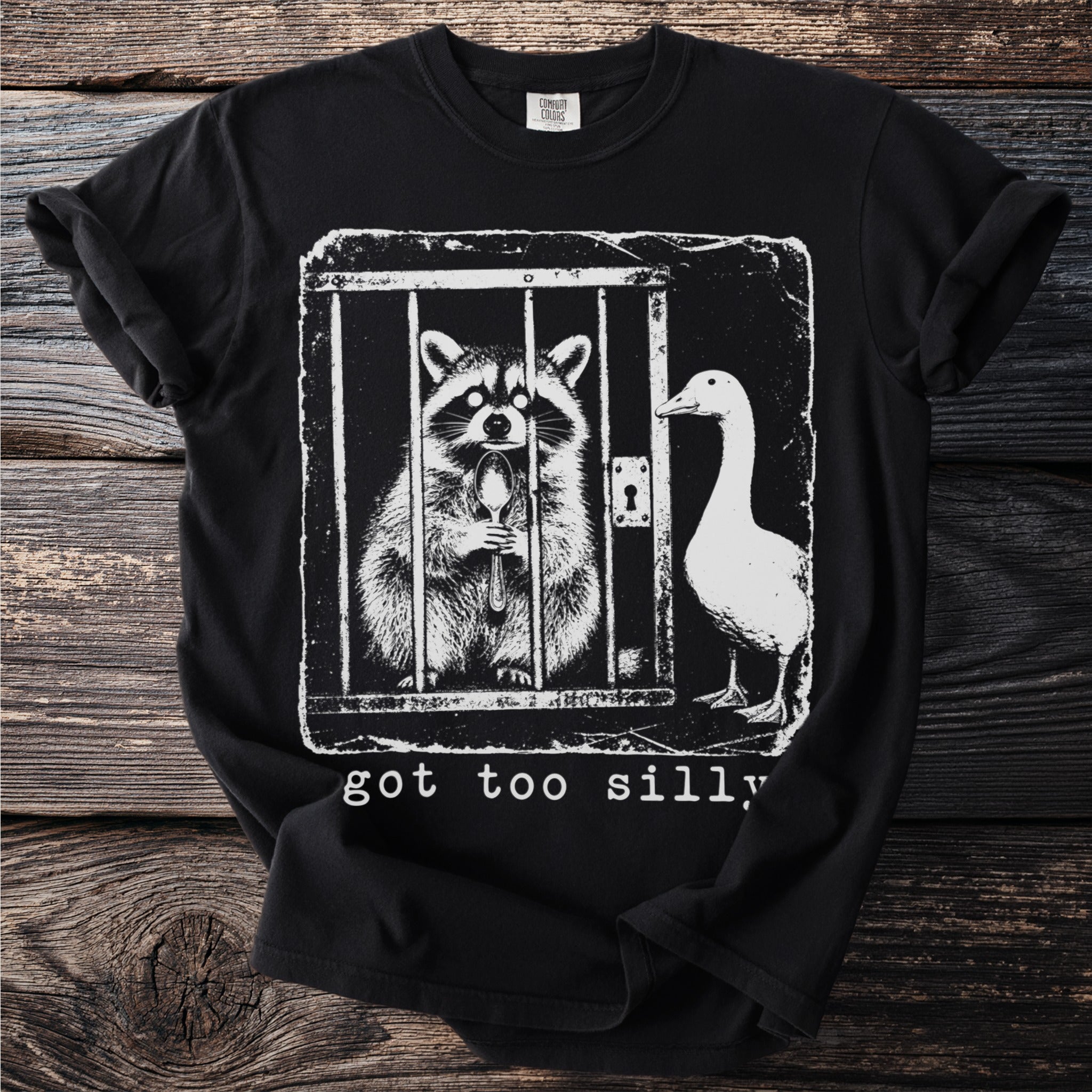 Black t-shirt with a graphic of a raccoon in a cage holding a spoon and a goose.