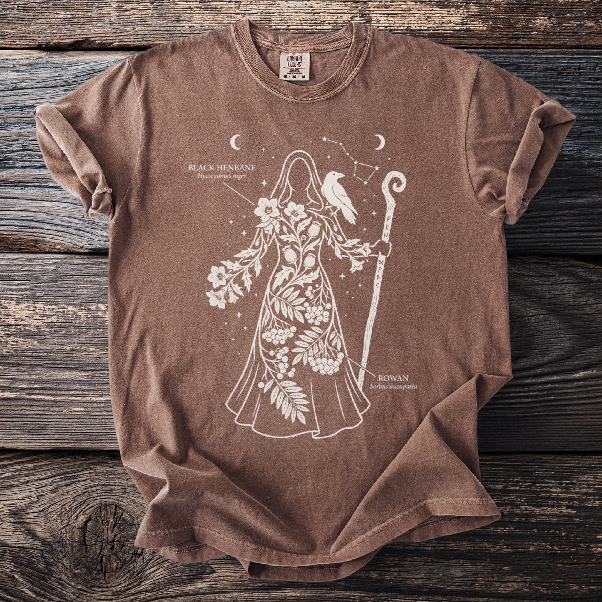 Brown t-shirt featuring a woman with flowers, a raven, and runes. Includes text "Black Henbane" and "Rowan".