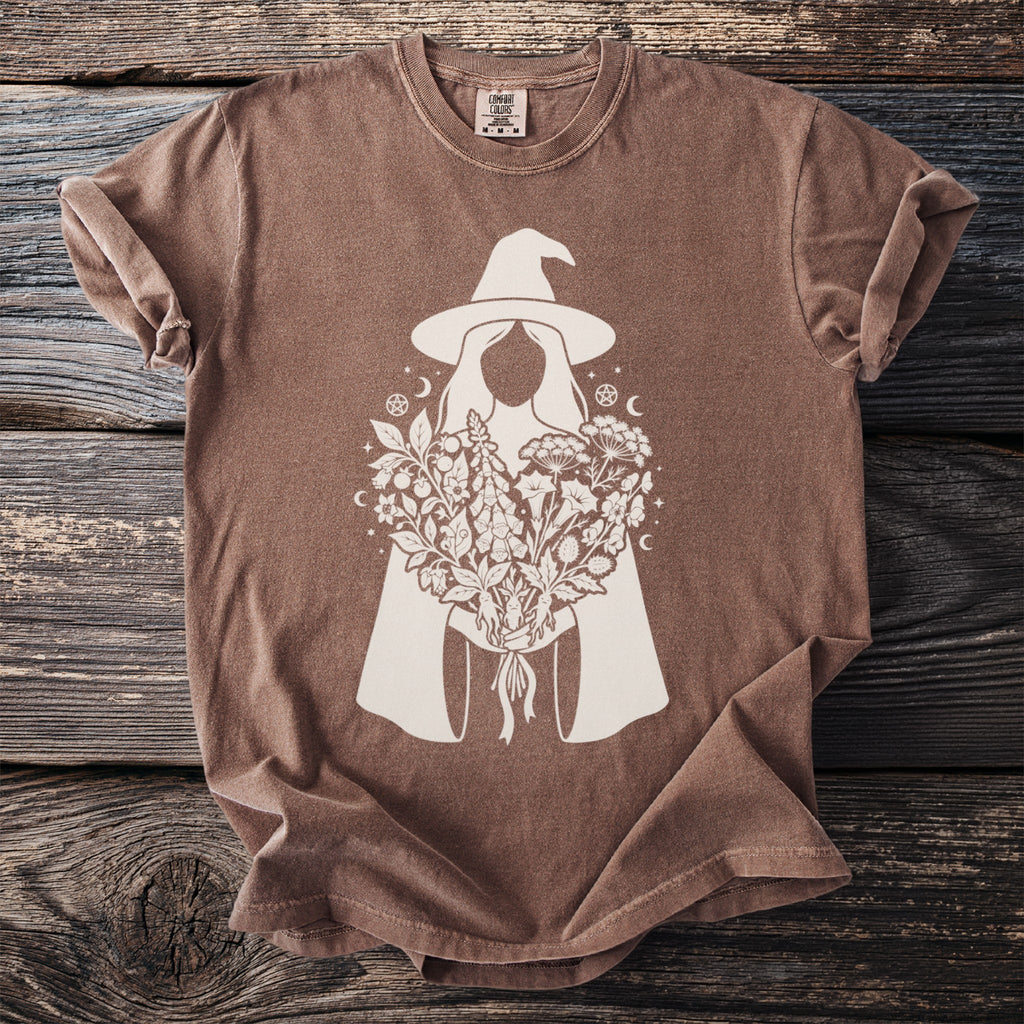 Brown t-shirt with white graphic of a witch holding a bouquet of herbs and flowers.