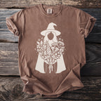 Brown t-shirt with white graphic of a witch holding a bouquet of herbs and flowers.