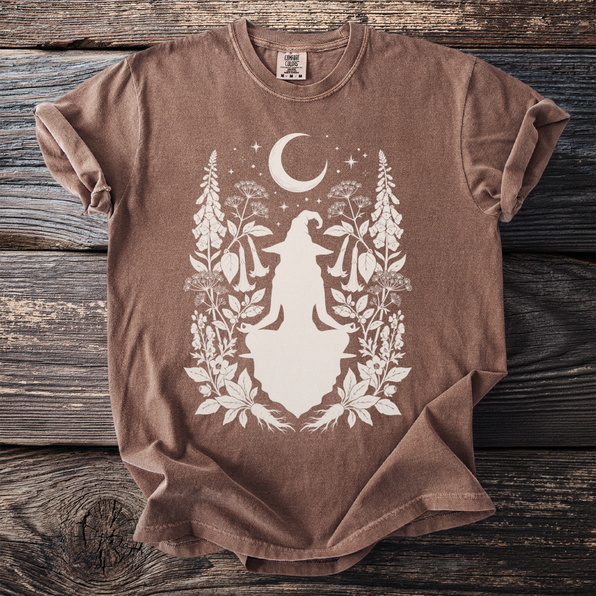 Brown t-shirt with a witch silhouette surrounded by crescent moon, stars, and botanical elements.