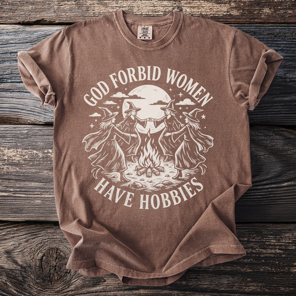 Brown t-shirt with graphic of witches around a campfire and text "GOD FORBID WOMEN HAVE HOBBIES".