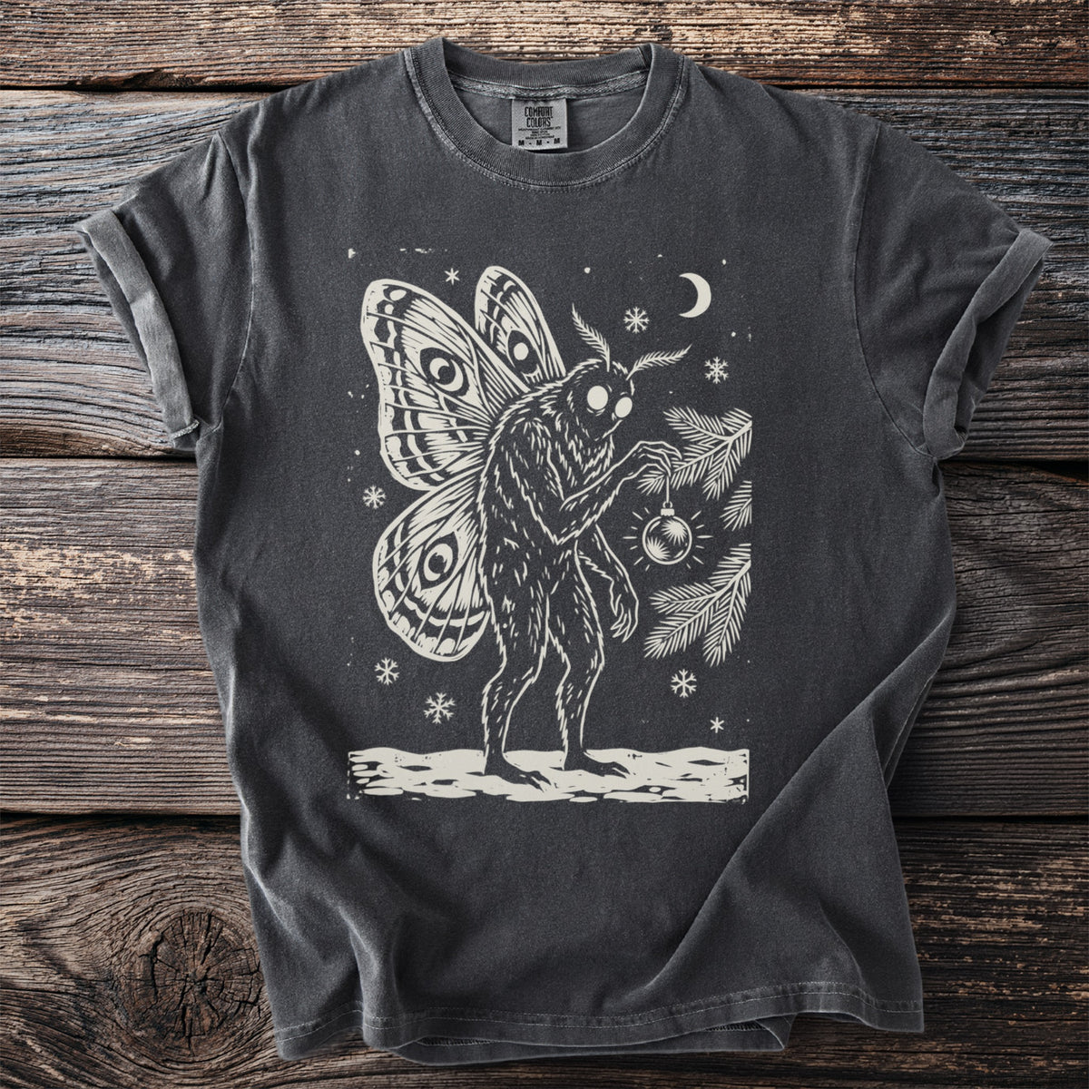 Pepper gray t-shirt with a cream folk-cut print: Mothman, eyespot wings open, hangs one glass ornament on a pine branch.
