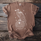 Brown t-shirt with white raven perched on ornamental ball, surrounded by snowflakes.