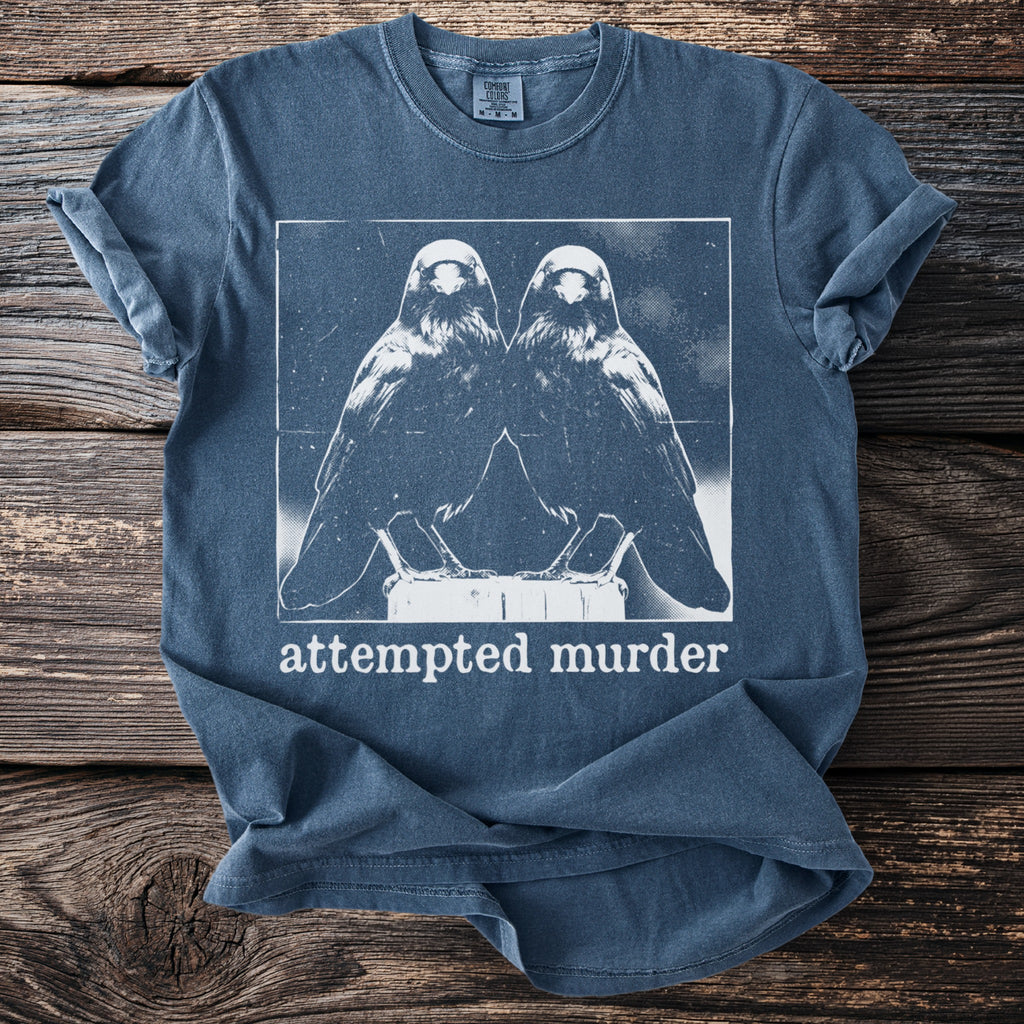 Blue t-shirt with a graphic of two mirrored birds and the text "attempted murder".
