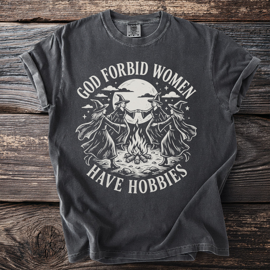 Dark t-shirt with four witches dancing around a campfire, text reads "God forbid women have hobbies".