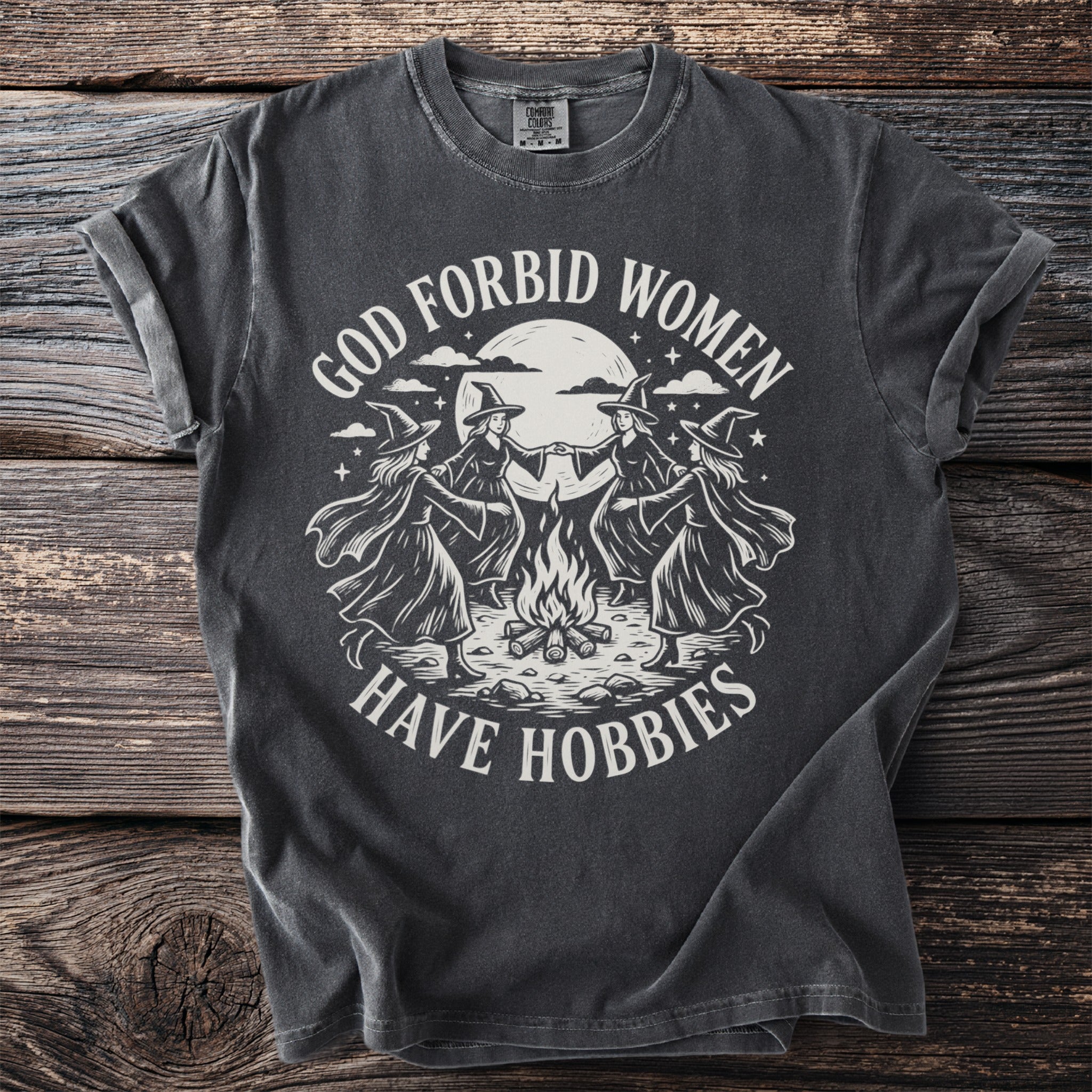 Dark t-shirt with four witches dancing around a campfire, text reads "God forbid women have hobbies".