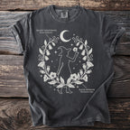 Dark t-shirt with white design of a witch silhouette, moon, stars, and botanical elements with text.