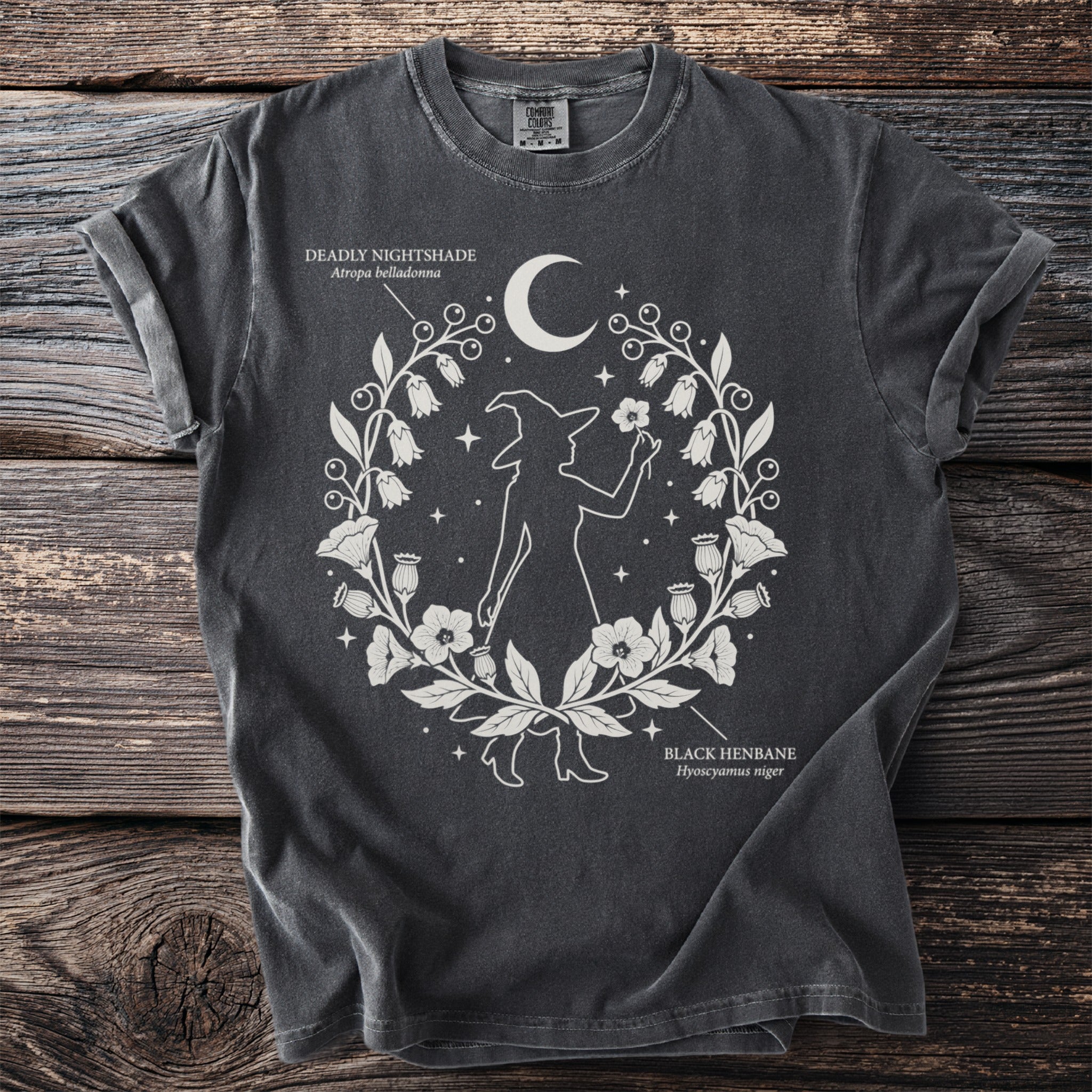 Dark t-shirt with white design of a witch silhouette, moon, stars, and botanical elements with text.