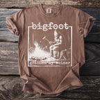 Brown t-shirt with Bigfoot sitting by a campfire, text reads "bigfoot grabbed my weiner".